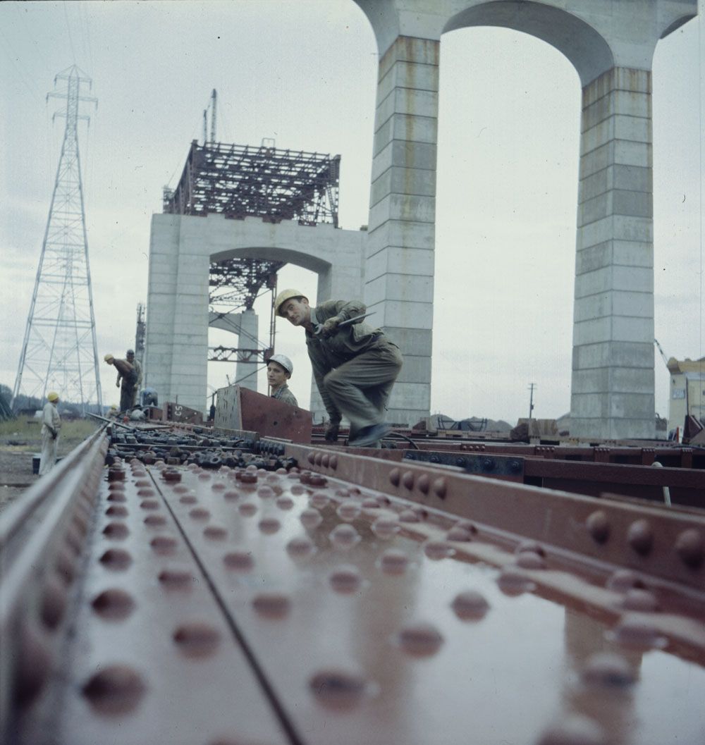 Construction workers climbing up on a section of the bridge, which is still on the ground. In the background are the large piers, which will