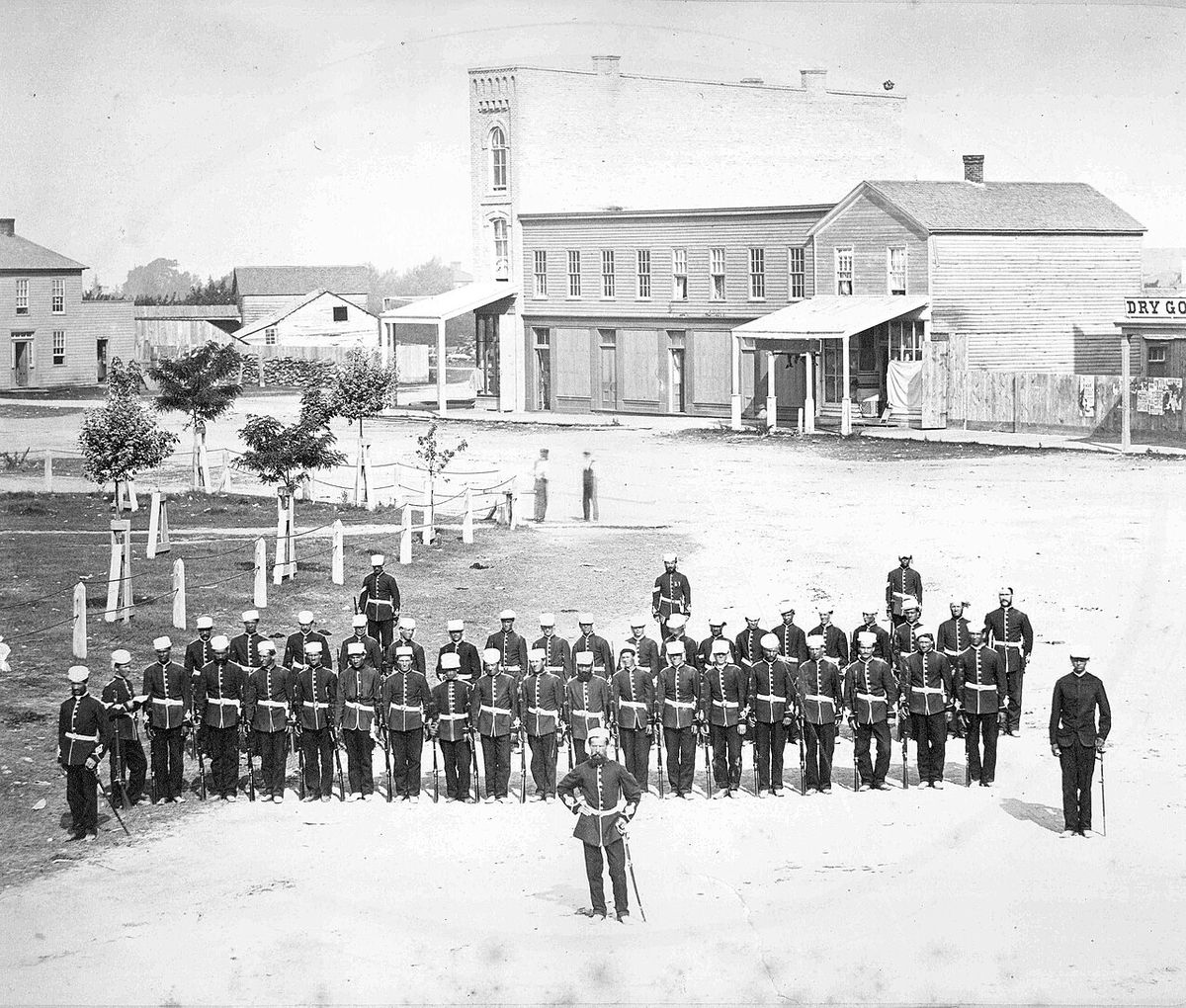 Huron County Militia Drilling on the Square, Goderich Ontario; sale posters on fence near "Dry Goods" store; names of stores not visible, po